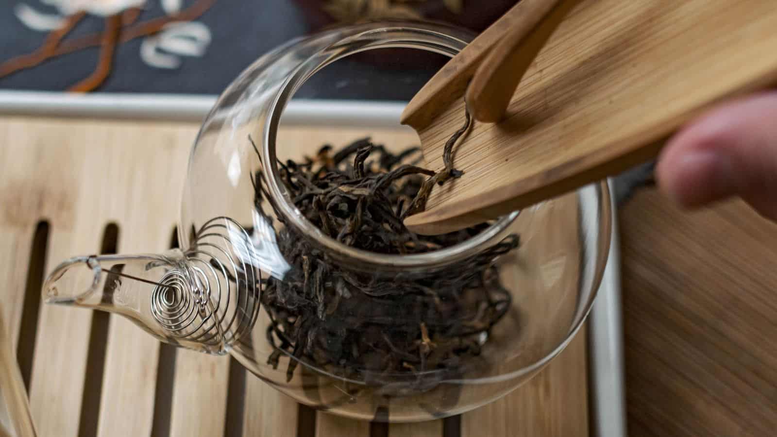 A hand uses wooden tongs to place dried tea leaves into a clear glass teapot. The teapot is set on a wooden tray with a spiral design visible on its spout. The background shows part of a decorated fabric.