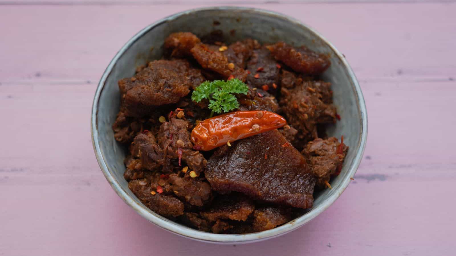 A bowl filled with cooked meat pieces seasoned with red chili flakes, garnished with a whole red chili pepper and a sprig of parsley. The bowl is placed on a pink wooden surface.