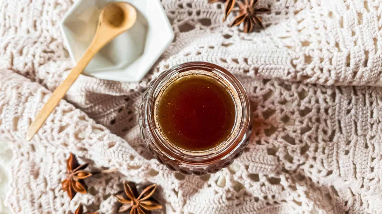 A glass jar filled with brown liquid sits on a crocheted cloth. Star anise and a wooden spoon on a white hexagonal dish are nearby, creating a rustic arrangement.