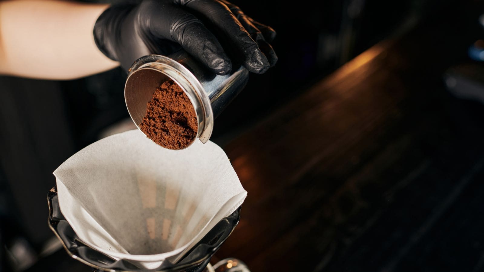 A person wearing black gloves pours ground coffee from a portafilter into a paper filter placed in a coffee dripper. The scene is set on a dark wooden surface.