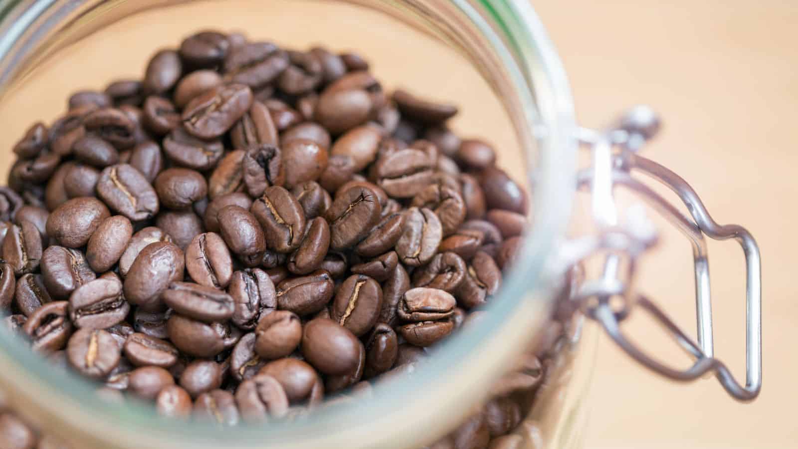 A glass jar filled with whole coffee beans in varying shades of brown. The jar is open, and the metal latch is partially visible on the right side. The surface beneath the jar is a light wood texture.