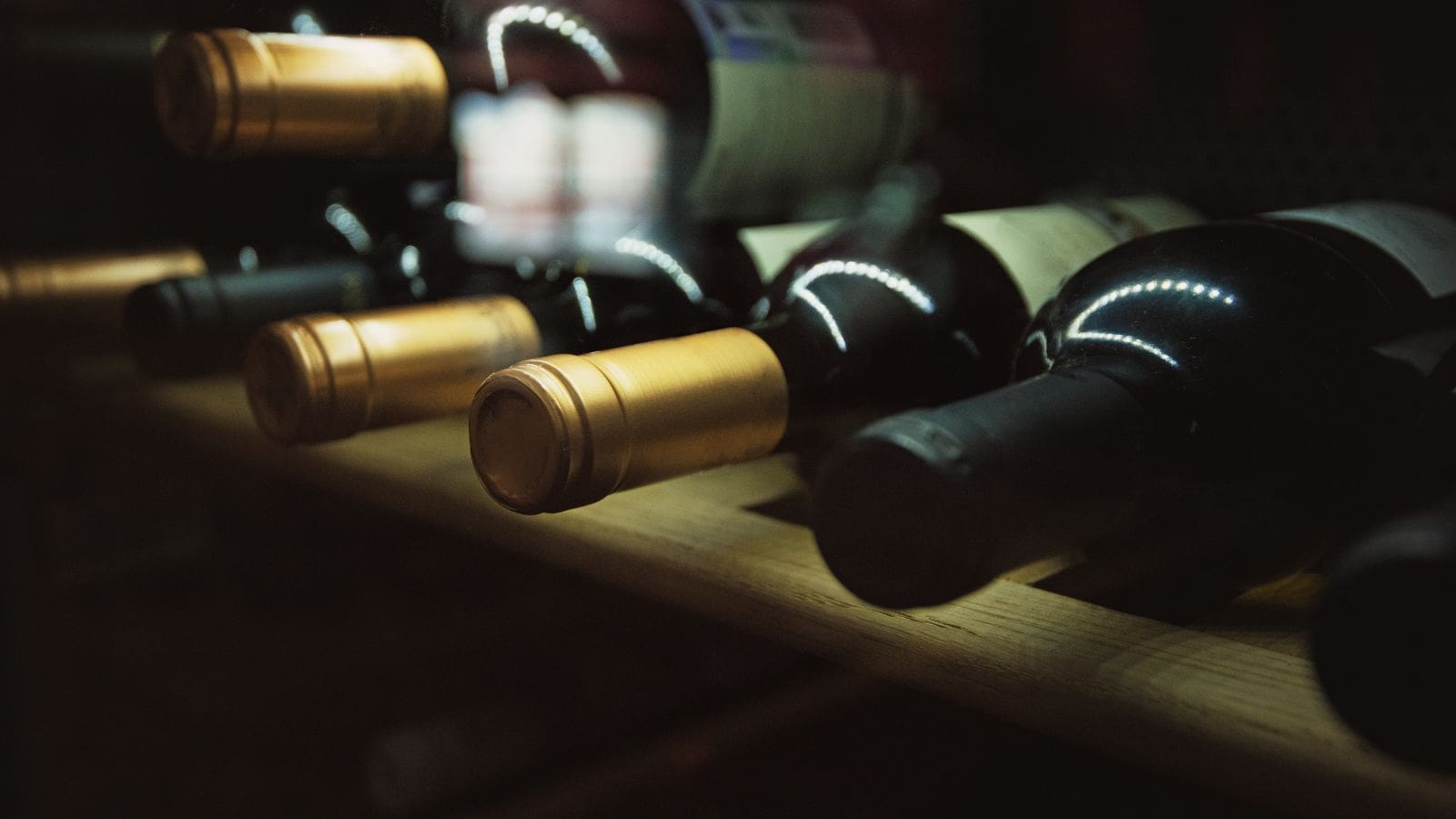 Bottles of wine lie horizontally on a wooden shelf. The focus is on the neck and corks of the bottles, which have gold and black foil coverings. The lighting is dim, creating a reflective sheen on the bottles.