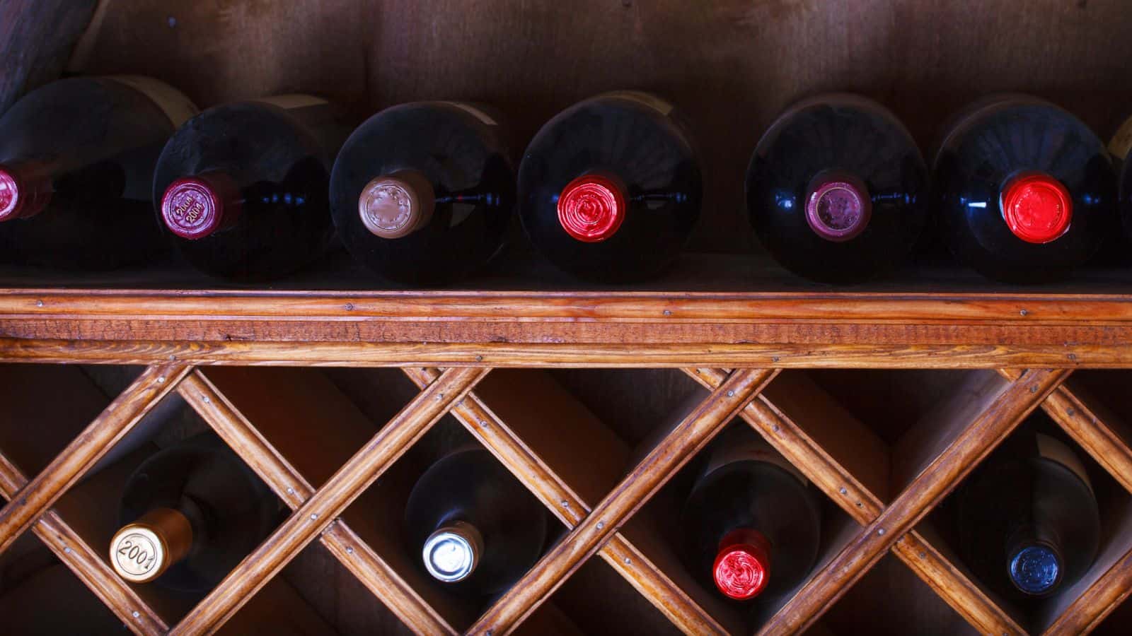 A wooden wine rack holds several bottles of wine. The bottles are stored horizontally, with various colored caps visible. Three more bottles are stored below in diagonal slots. The rack has a rustic appearance, and the background is a wooden surface.