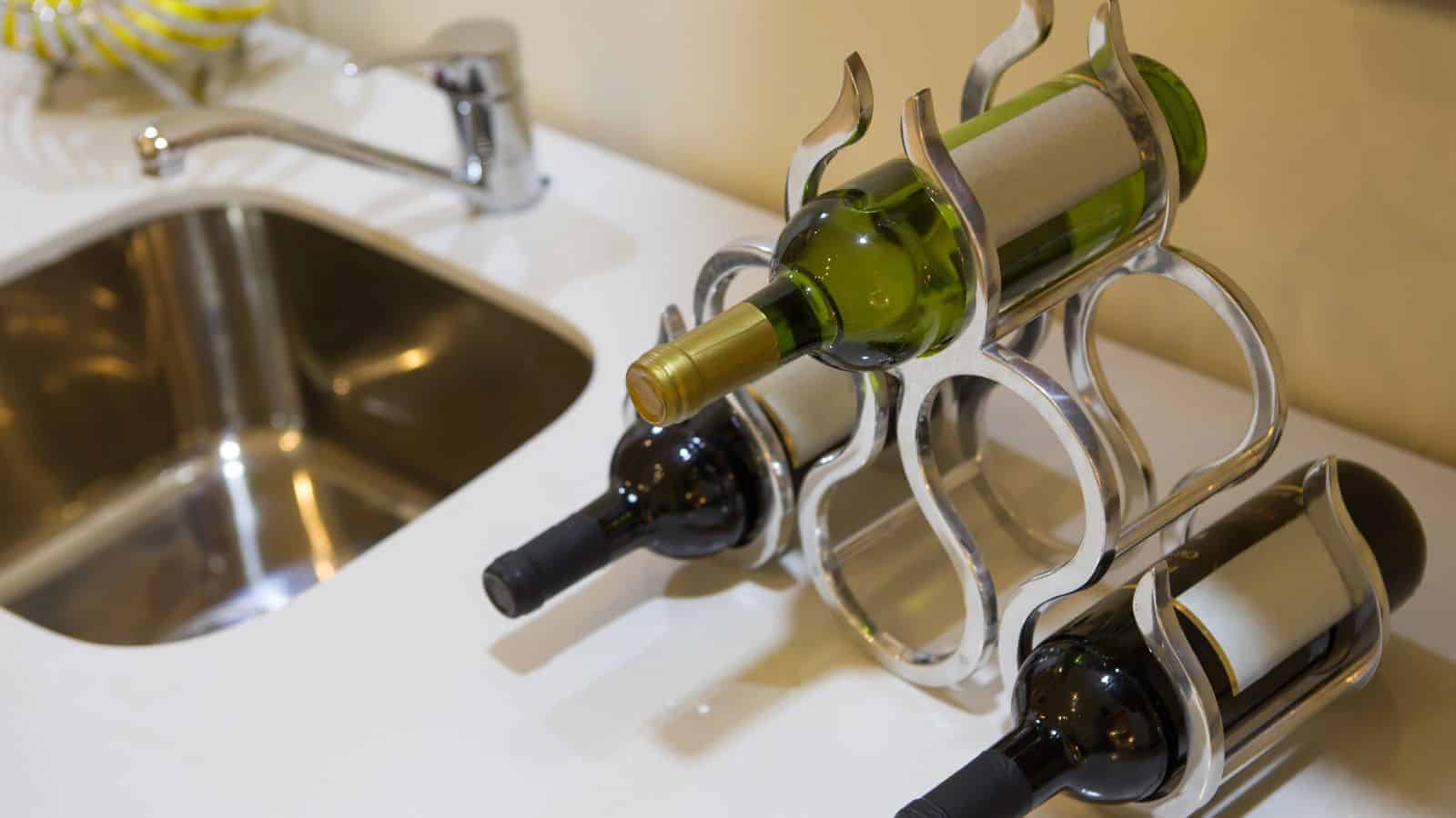 Three wine bottles, one white and two red, are stored horizontally in a silver, decorative wine rack on a white countertop. A stainless steel sink and faucet are visible to the left.
