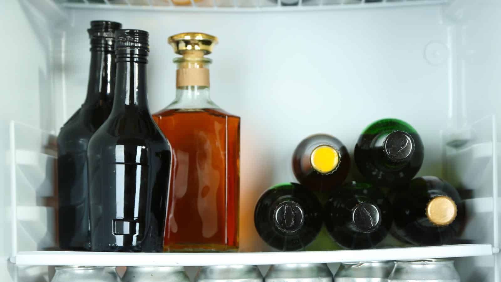 A refrigerator shelf containing three dark glass bottles with long necks, one square glass bottle with amber liquid and a gold cap, and five dark green glass bottles with yellow caps lying on their sides. Below are aluminum cans.