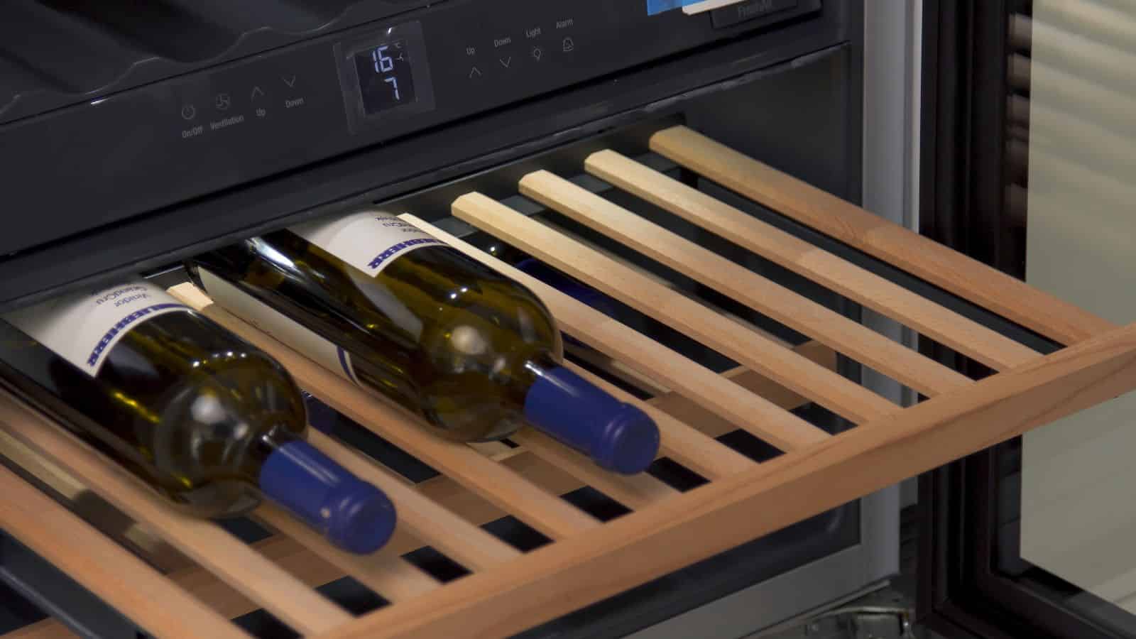 A wine refrigerator with a wooden rack holding three bottles of wine with blue caps. The display shows a temperature of 16.7 degrees. The door is partially open, revealing the slatted shelf design.