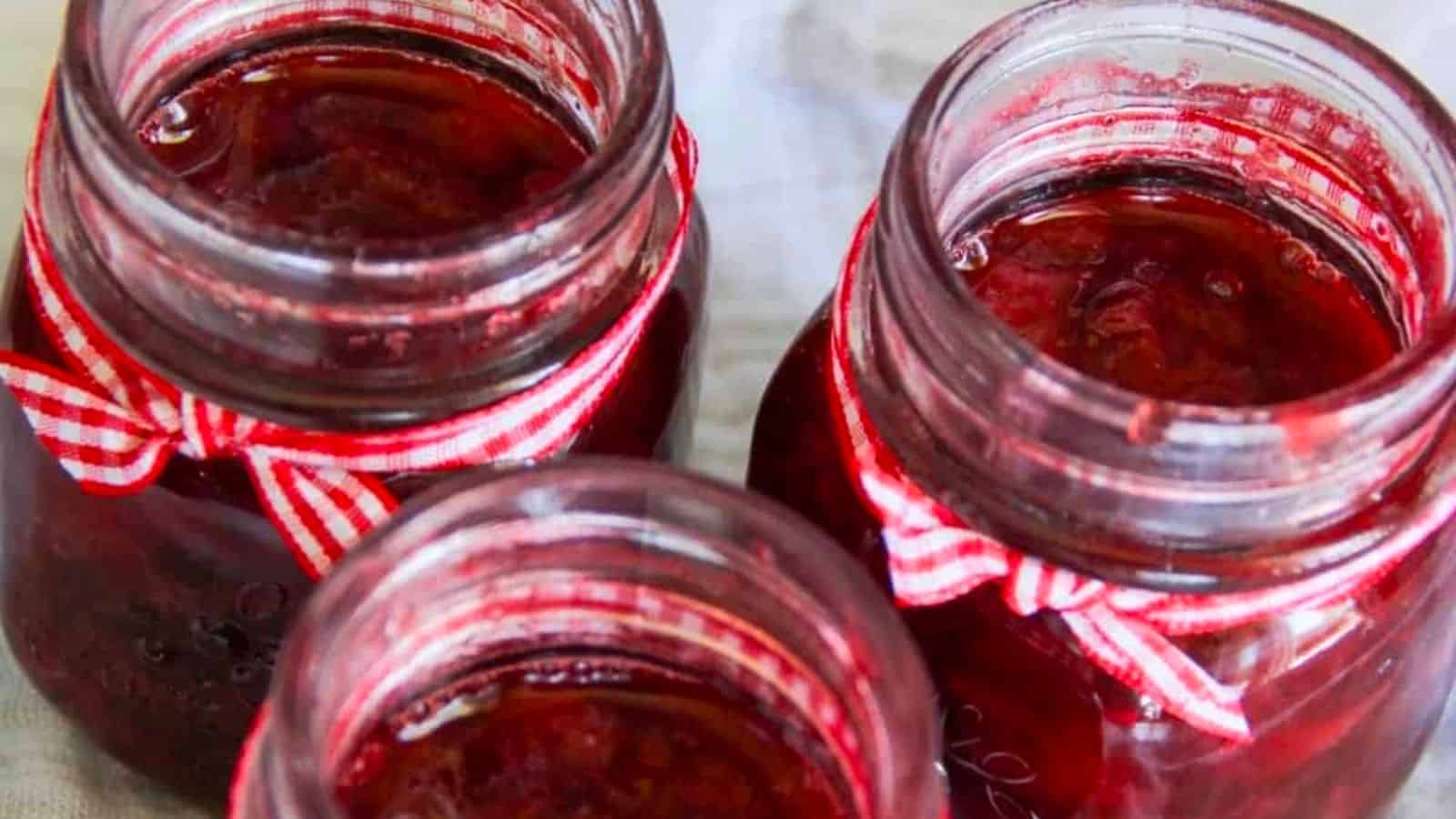 Three glass jars filled with red jam are arranged closely together. Each jar is adorned with a red and white checkered ribbon tied around its neck. The jars are open, revealing the thick consistency of the jam inside.