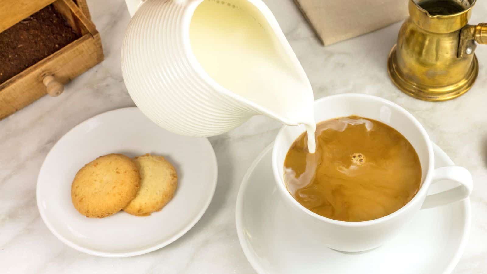 A pot of milk is being poured into a cup of coffee on a table. Next to the cup are two round cookies on a white plate. A brass container and a wooden box are in the background.