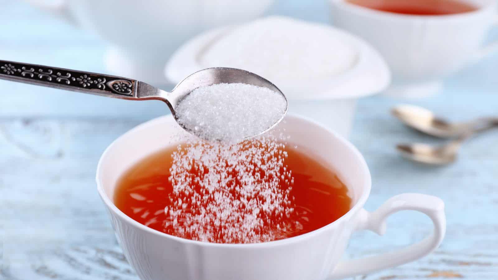A spoon is pouring sugar into a cup of tea. Two white cups filled with tea are visible in the background, along with a sugar bowl and two spoons on a light blue wooden surface.