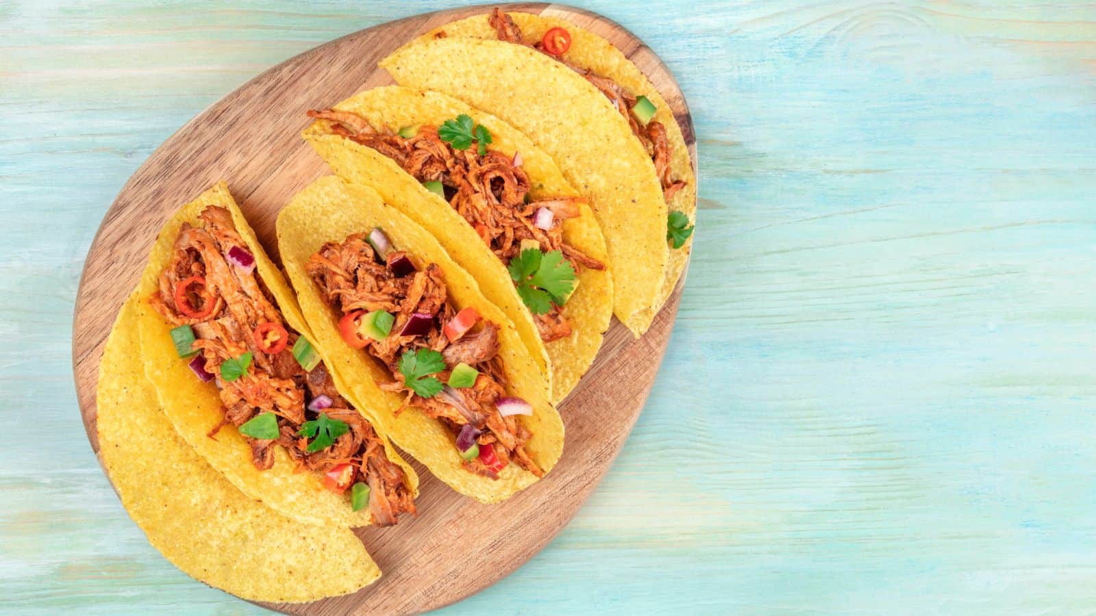Three hard shell tacos filled with shredded meat, sliced red onions, red chili peppers, and cilantro are arranged on a wooden board. The background is a textured, light blue surface.
