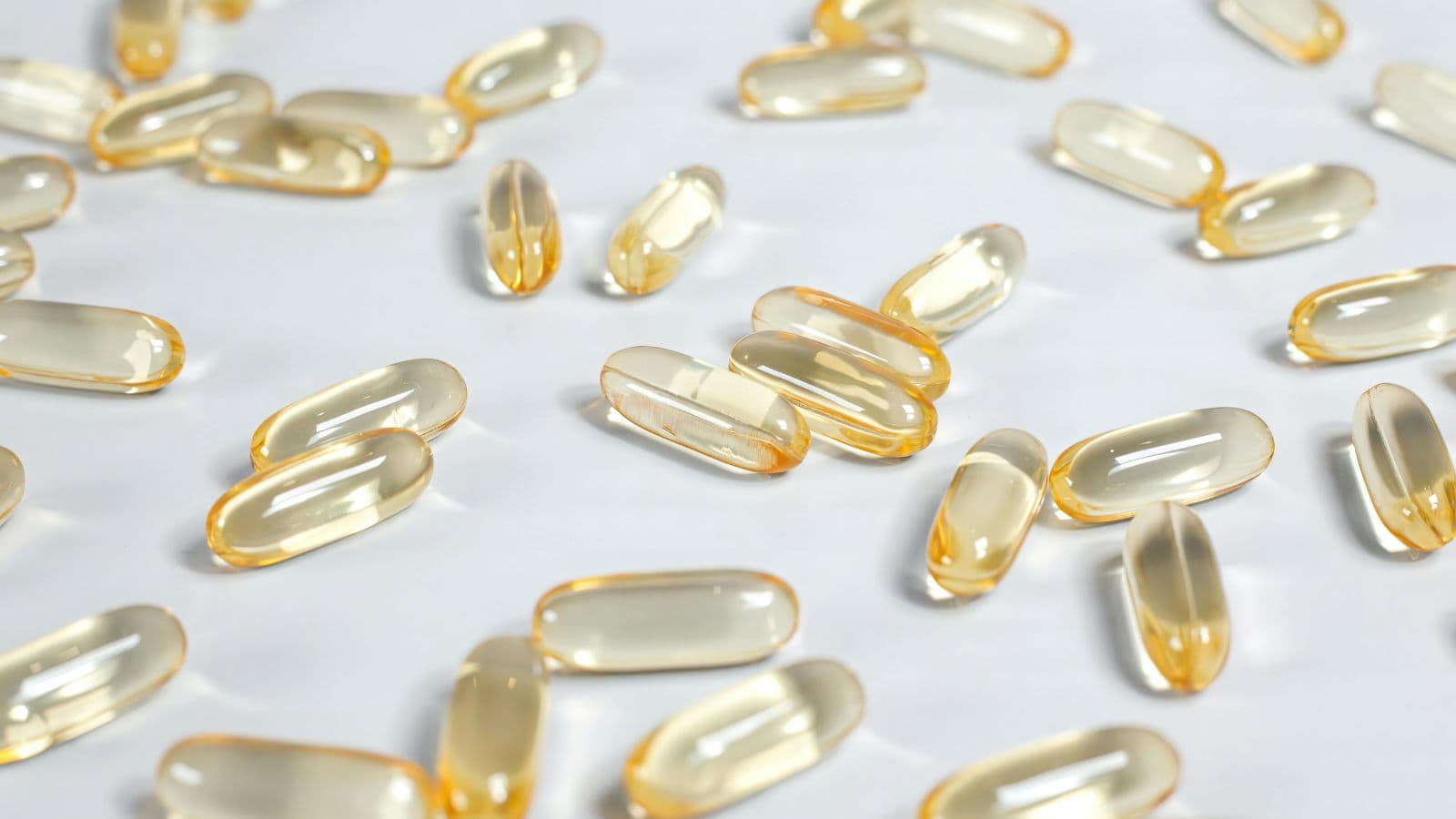 A scattered collection of clear, oval-shaped gel capsules with a yellow tint on a white surface. The capsules vary in their orientation, with some laying flat and others on their sides.