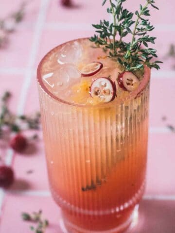 A tall, ribbed glass filled with an orange and pink gradient cocktail, topped with ice cubes, cranberries, and a sprig of thyme. The glass is on a pink tiled surface with scattered cranberries and thyme sprigs.