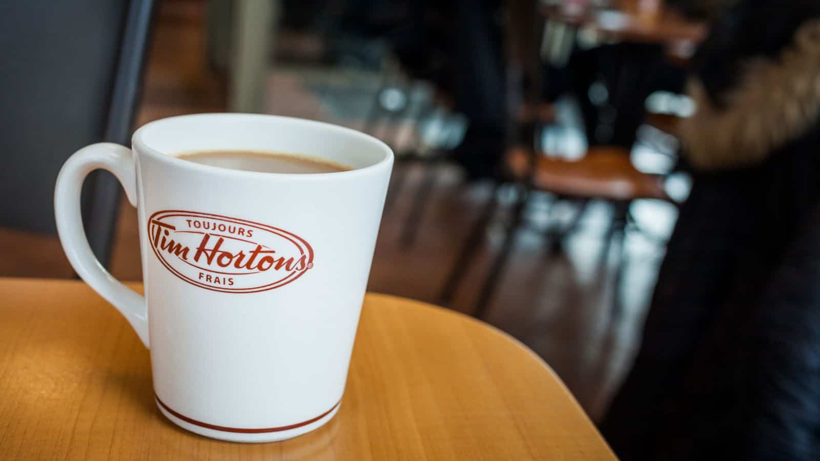 A white mug with the Tim Hortons logo filled with coffee sits on a wooden table in a cafe setting. The background shows blurred chairs and a person wearing a dark coat.