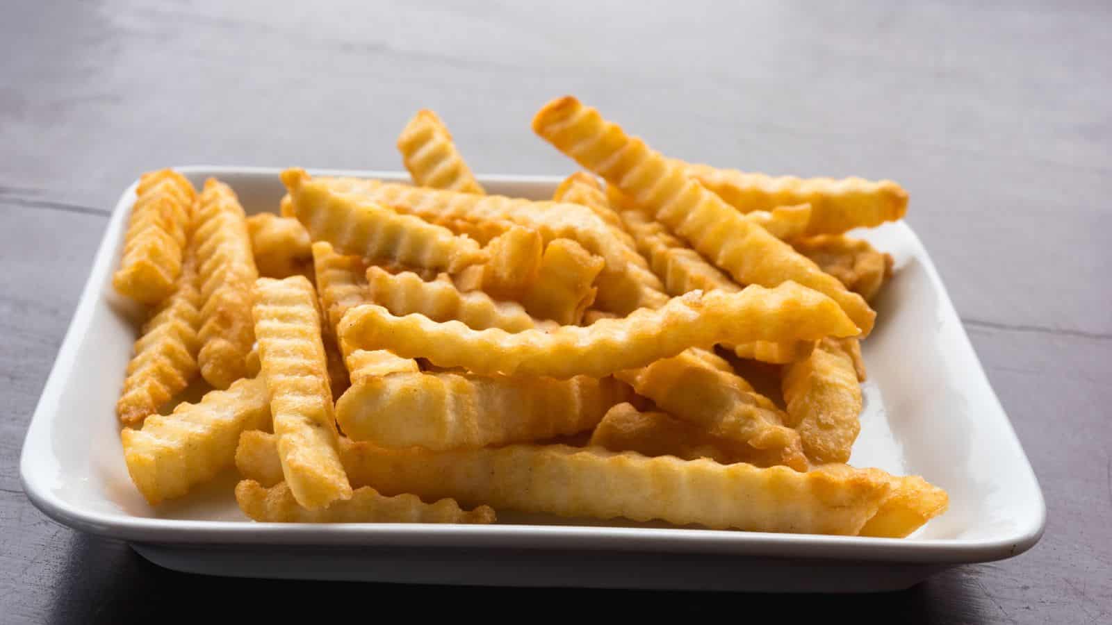 A white rectangular plate filled with crinkle-cut French fries. The fries are golden brown and appear crispy. The background is a dark, smooth surface.