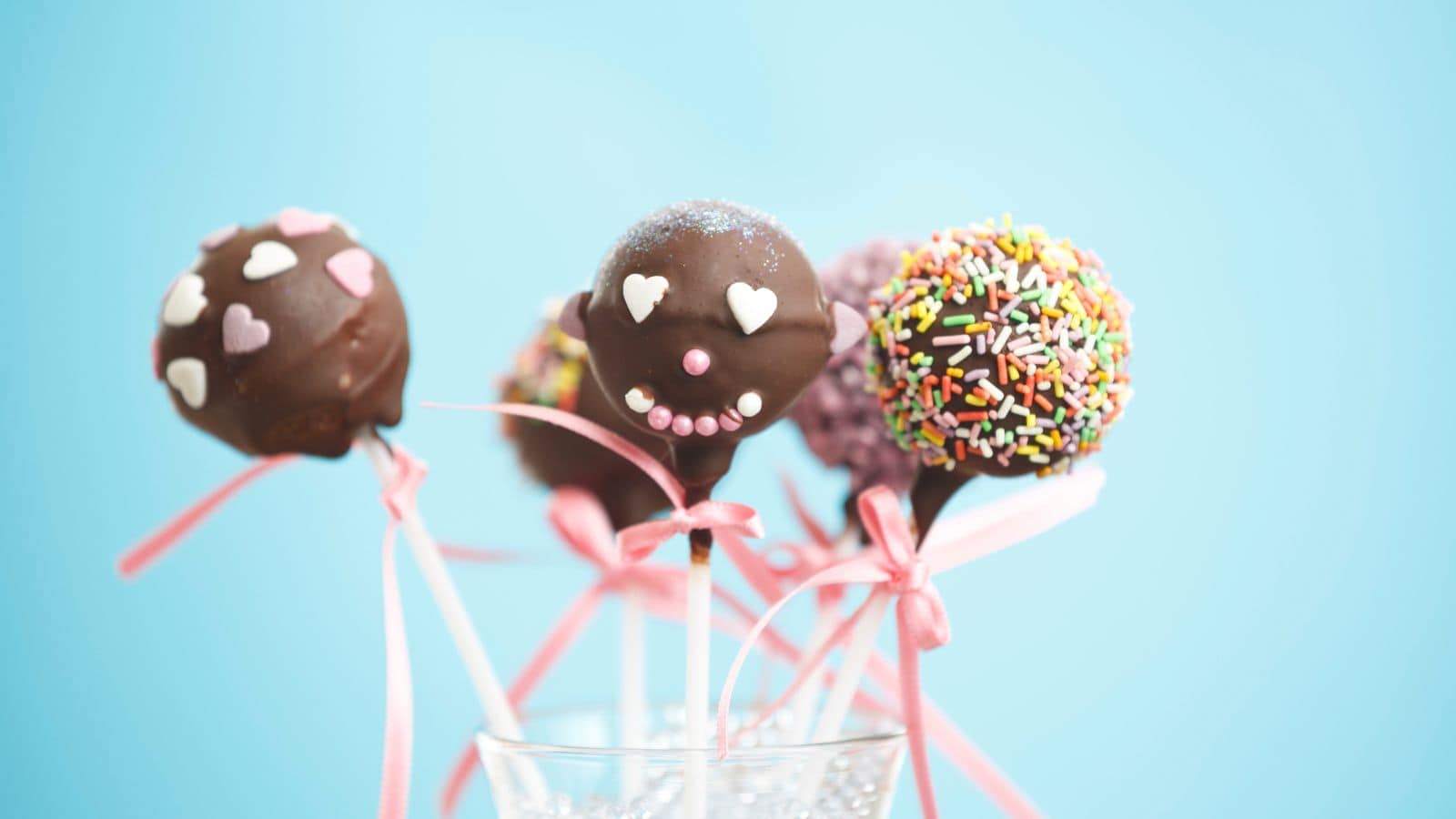 Four cake pops with various chocolate coatings and decorations including heart-shaped sprinkles, a smiley face, and colorful sprinkles are displayed in a glass cup against a light blue background. Each cake pop is tied with a pink ribbon.