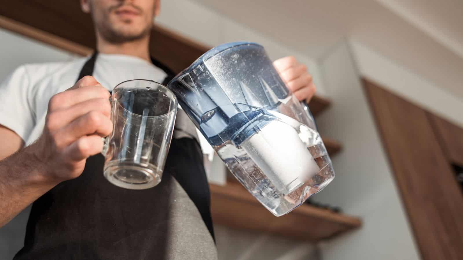 A person wearing an apron holds a clear glass mug in one hand and a water filter pitcher in the other. They are standing in a kitchen with wooden shelves visible in the background.