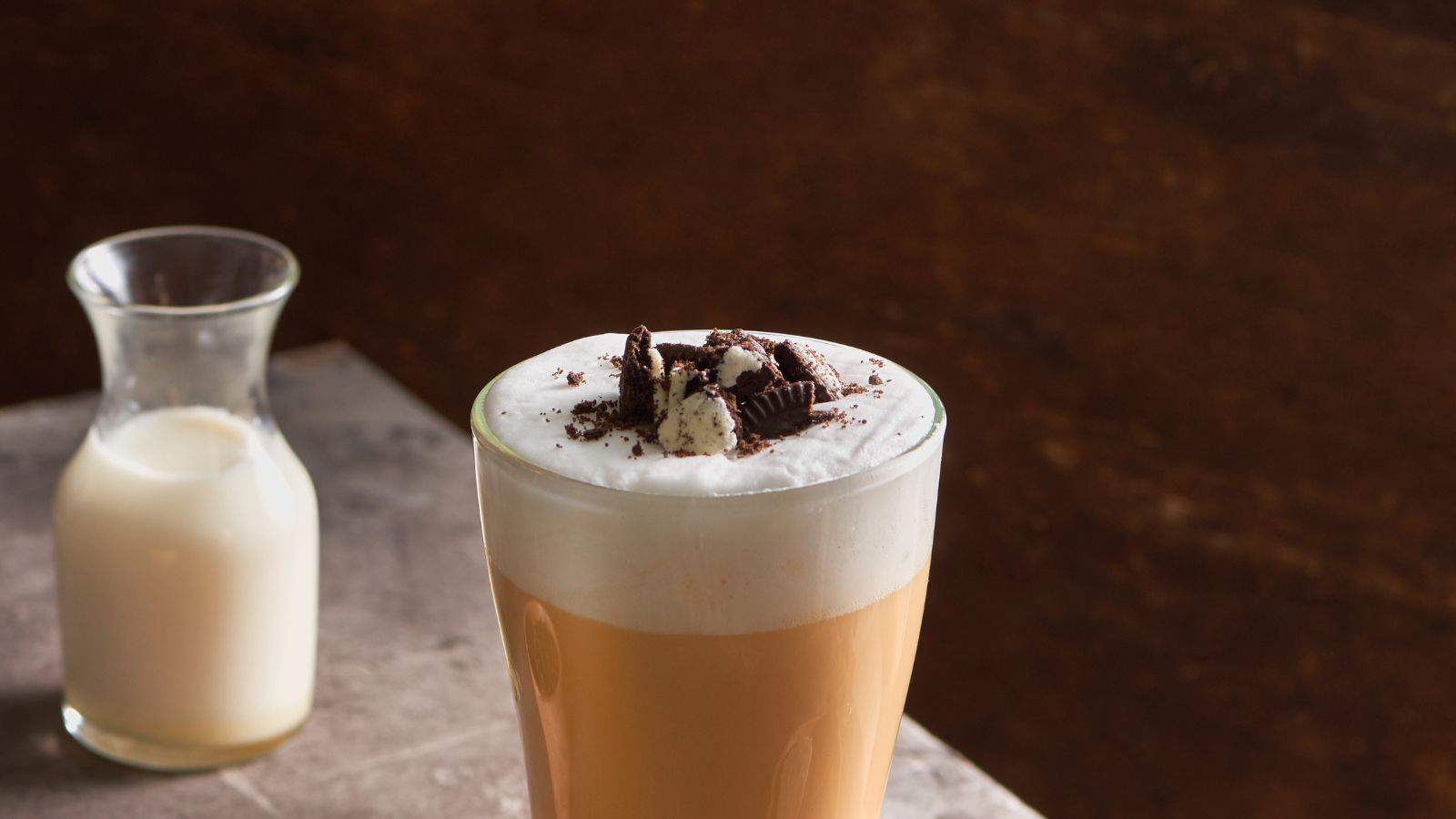 A glass of iced coffee topped with whipped cream and crushed cookies sits on a stone counter. In the background, there is a small glass jug filled with milk. The surface has a rustic texture, and the background is dark brown.