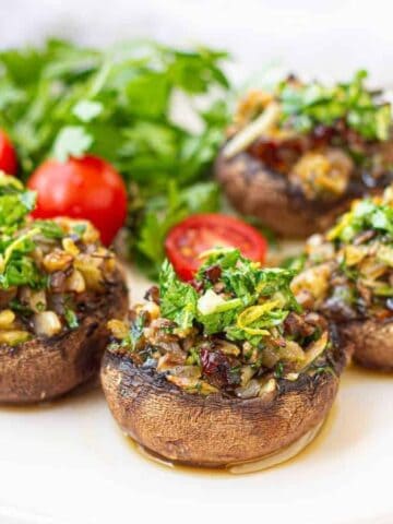 Four stuffed mushrooms are on a white plate, topped with herbs and chopped vegetables. Accompanied by cherry tomatoes and parsley. The background is blurred, with a white surface and soft lighting.