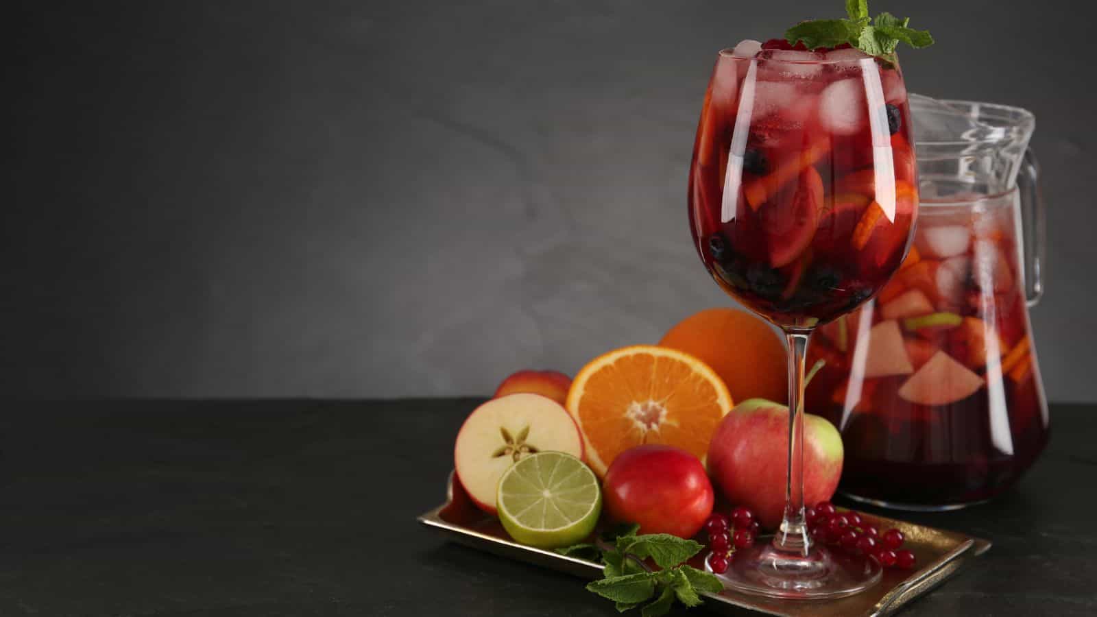 A glass and a pitcher of red sangria with ice and fruit slices sit on a dark surface. In front, an assortment of fresh fruits including apples, oranges, lime, and mint leaves are arranged on a tray. The background is plain and dark.