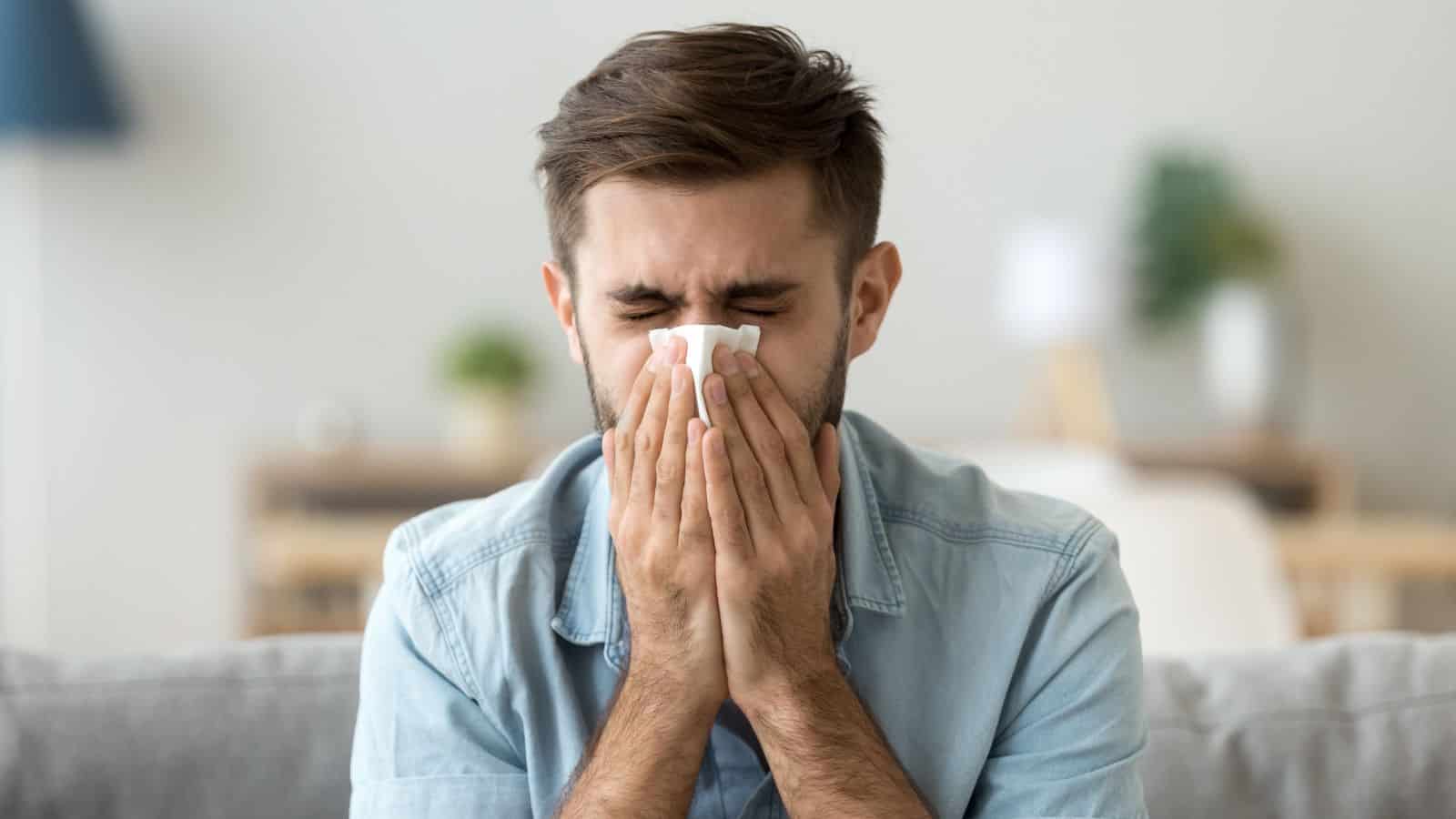 A man in a denim shirt sits on a couch holding a tissue to his nose with his eyes closed, appearing to sneeze. The background shows a blurred interior with a lamp and plants.