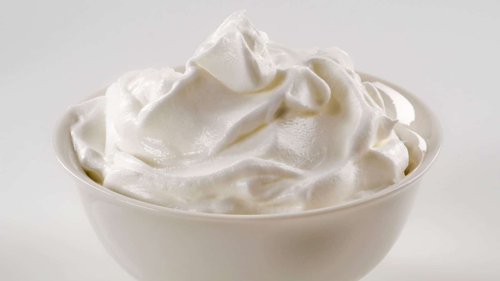 A white bowl filled with a mound of smooth, creamy whipped cream. The texture appears light and fluffy, with gentle peaks forming on the surface. The background is a plain, light gray color.