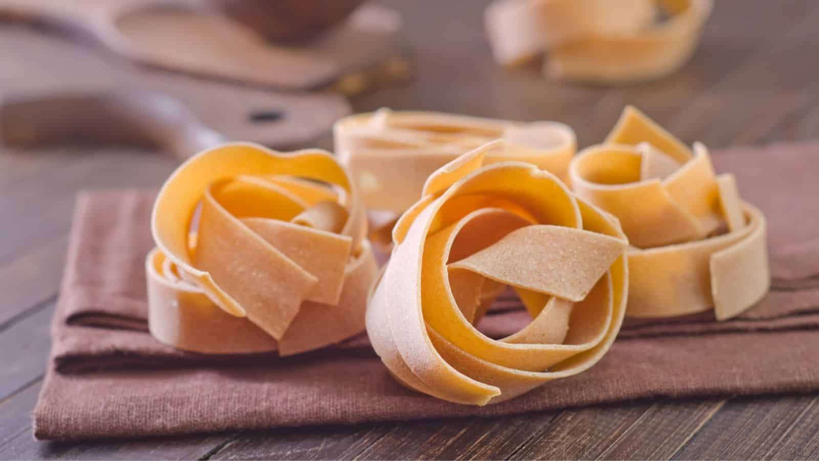 Rolled nests of uncooked tagliatelle pasta sit on a wooden surface atop a brown cloth. A blurred kitchen utensil is in the background. The pasta is light yellow and neatly arranged, suggesting preparation for cooking.