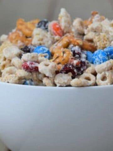 A white bowl filled with a snack mix containing pretzels, chocolate candies, cereal pieces, and nuts sits on a wooden surface. The mix has a variety of textures and colors, including blue and orange candies.