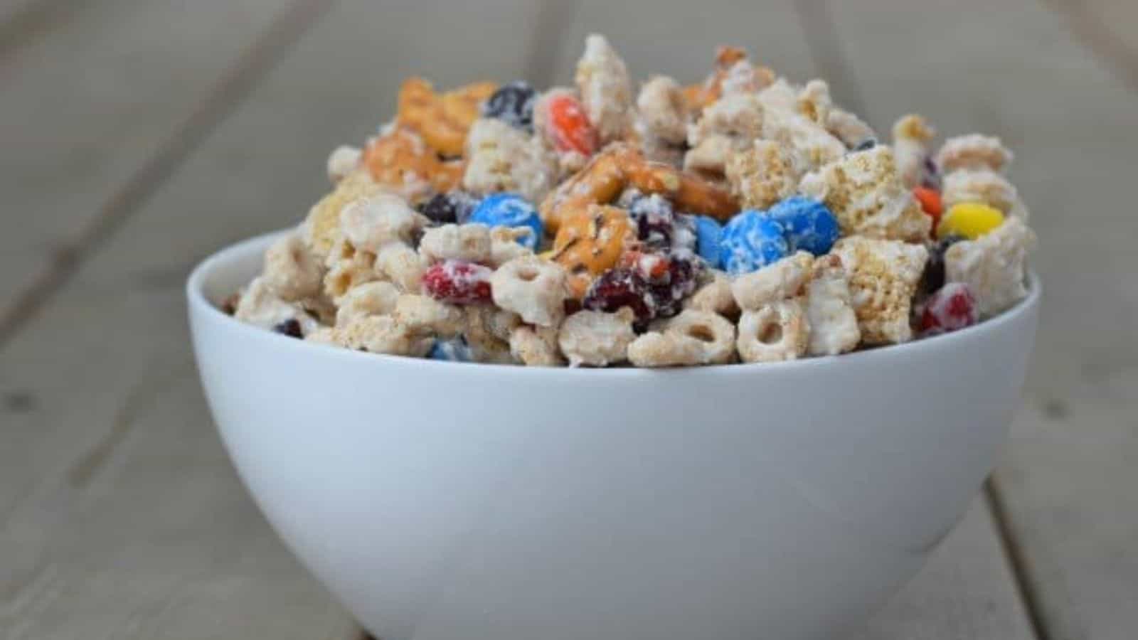 Image shows white chocolate party mix piled into a white bowl on a wooden table.