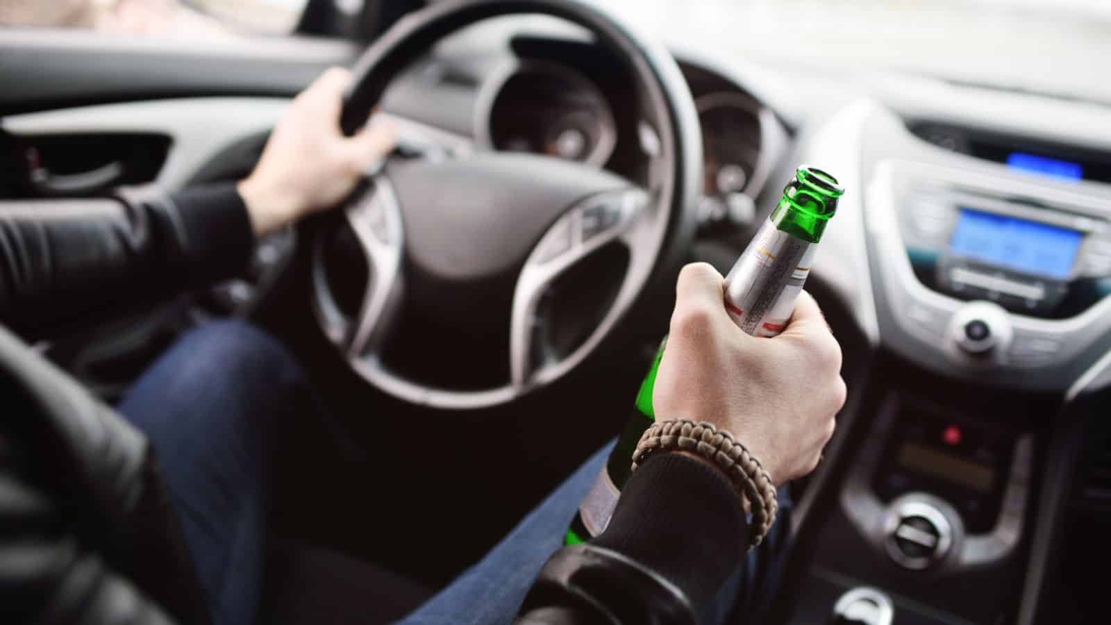 A person is holding a green glass bottle in their right hand while steering a car with their left hand. The car's interior, including the steering wheel and dashboard, is visible. The person is wearing a dark jacket and a bracelet.