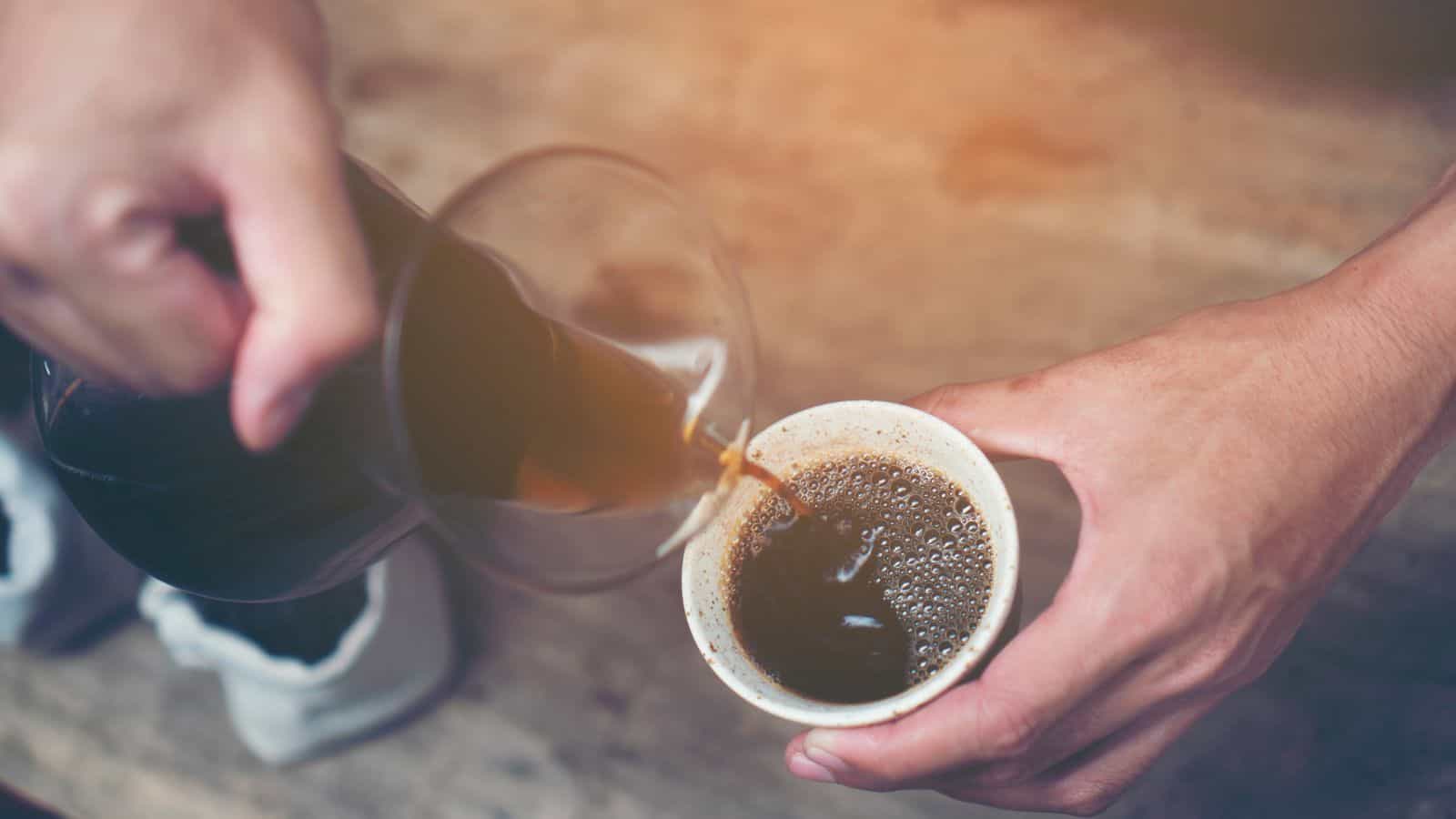 A person pours coffee from a glass carafe into a white paper cup. The background features a wooden surface, and another hand holds the cup steady. The coffee appears freshly brewed with visible bubbles on the surface.