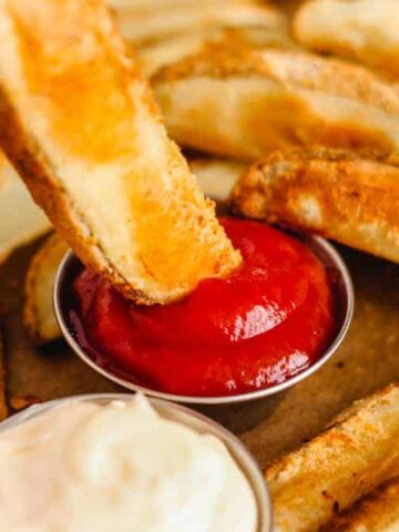 Close-up of a hand dipping a wedge-shaped potato fry into a small metal cup of ketchup. Several other potato wedges are scattered on a surface. Another cup with a creamy white sauce is also present.