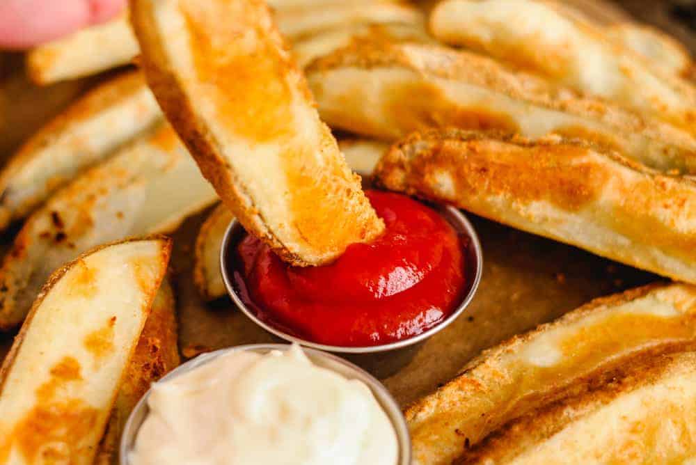 Close-up of a hand dipping a wedge-shaped potato fry into a small metal cup of ketchup. Several other potato wedges are scattered on a surface. Another cup with a creamy white sauce is also present.