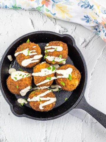 A black skillet holds six breaded and fried balls topped with a creamy white sauce and garnished with chopped herbs. A floral-patterned cloth is partially visible to the right on a light textured surface.