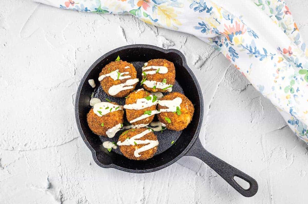 A black skillet holds six breaded and fried balls topped with a creamy white sauce and garnished with chopped herbs. A floral-patterned cloth is partially visible to the right on a light textured surface.