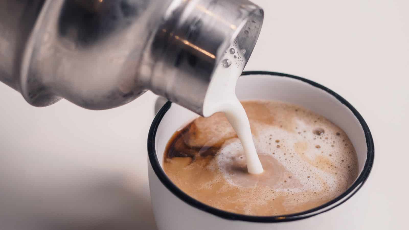 A metal container is pouring foamed milk into a white coffee cup with a black rim, creating a swirl pattern on the surface of the coffee. The background is plain and beige.