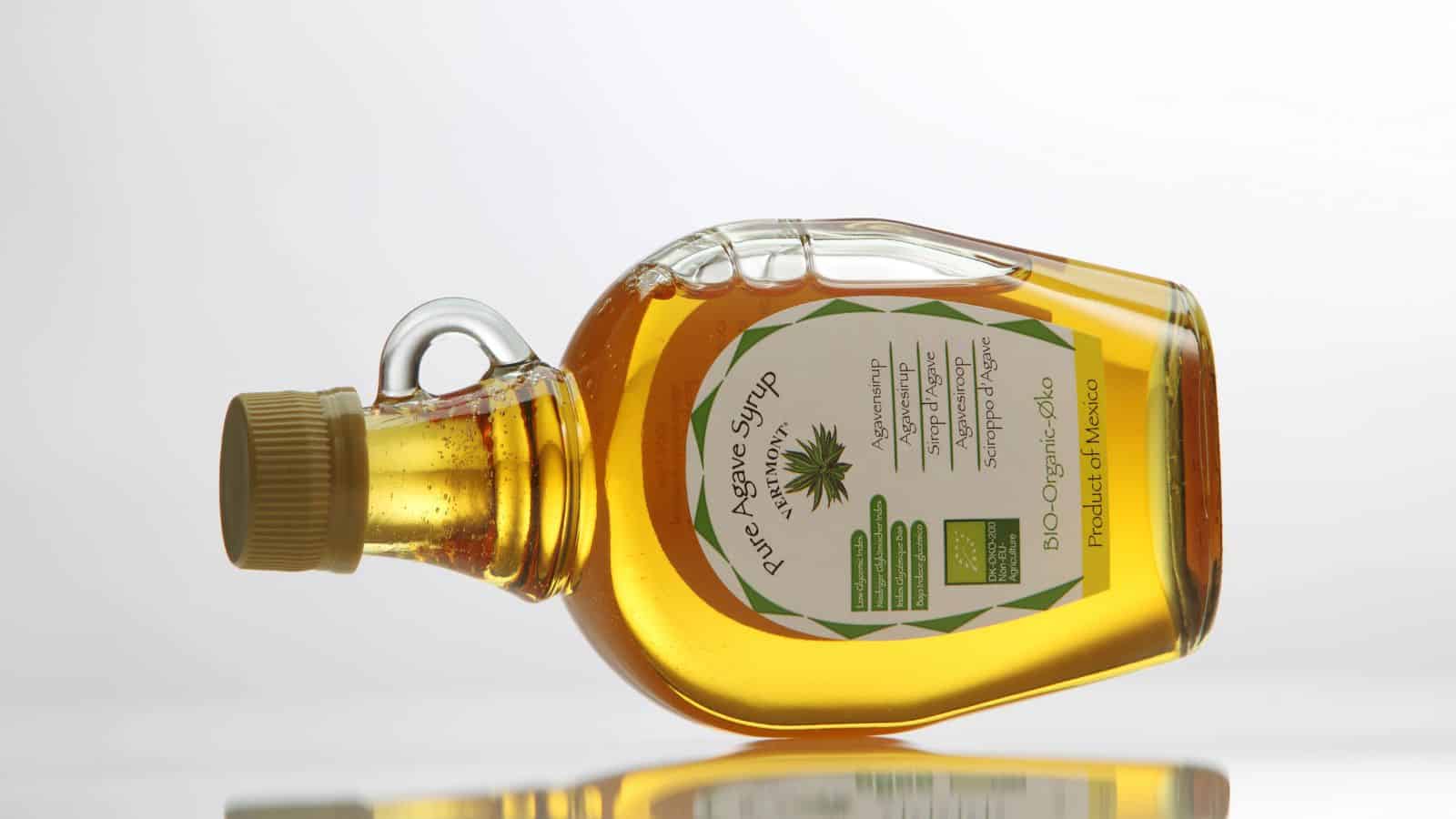 A clear bottle of pure agave syrup is placed on its side against a plain background. The bottle has a green and white label with text and an image of an agave plant. The syrup inside is amber-colored.