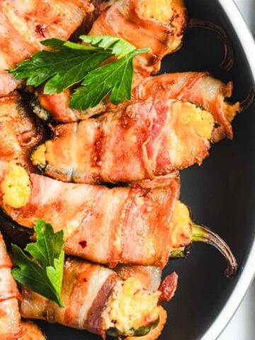 Bacon-wrapped jalapeño poppers in a black skillet, garnished with parsley. A grater and shredded cheese are on a wooden cutting board beside the skillet.