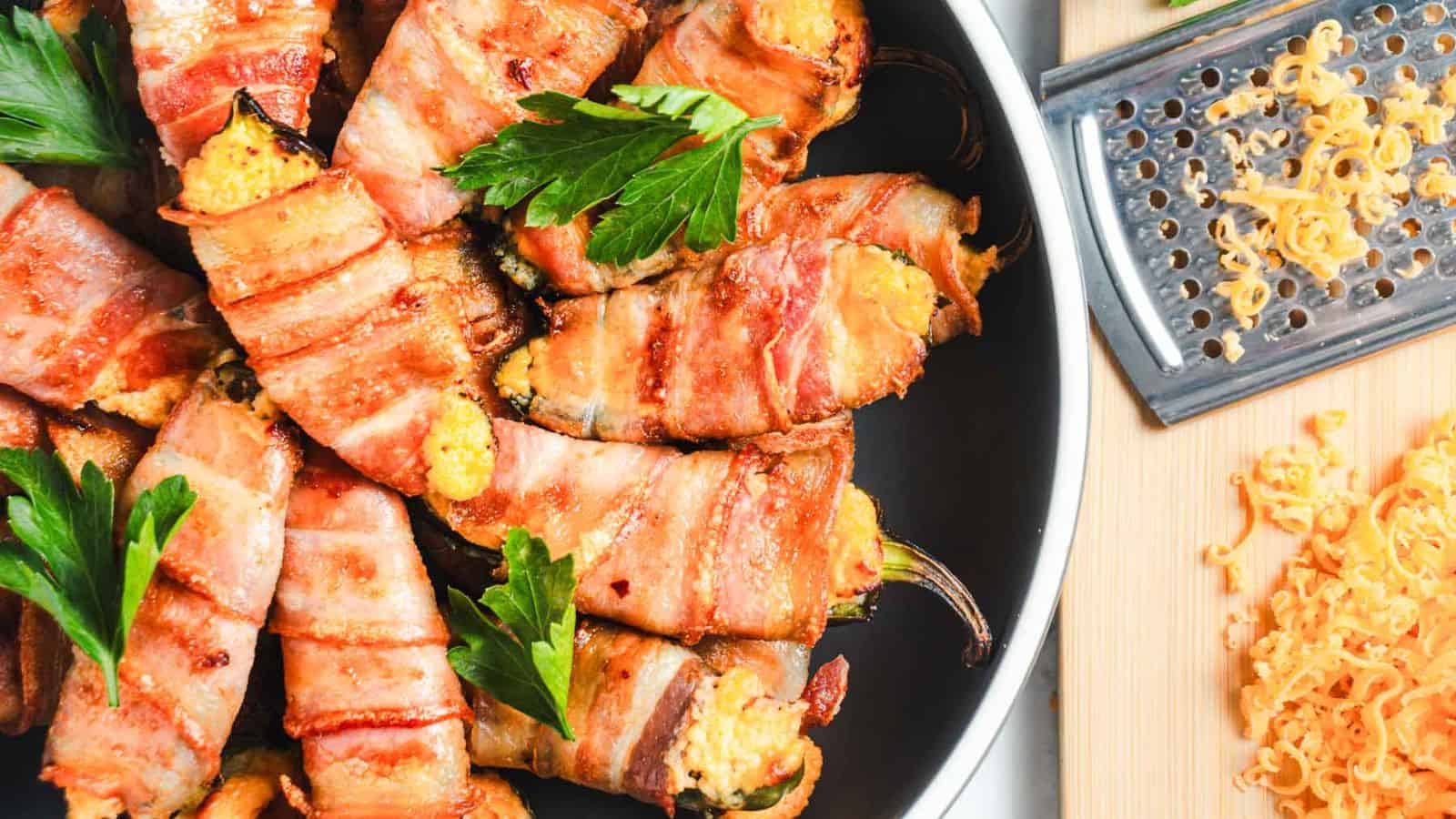 Bacon-wrapped jalape&ntilde;o poppers in a black skillet, garnished with parsley. A grater and shredded cheese are on a wooden cutting board beside the skillet.
