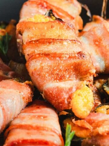 Close-up of bacon-wrapped jalapeño poppers in a pan, garnished with parsley leaves. The poppers are crispy and golden brown, with cheese oozing out from the ends.