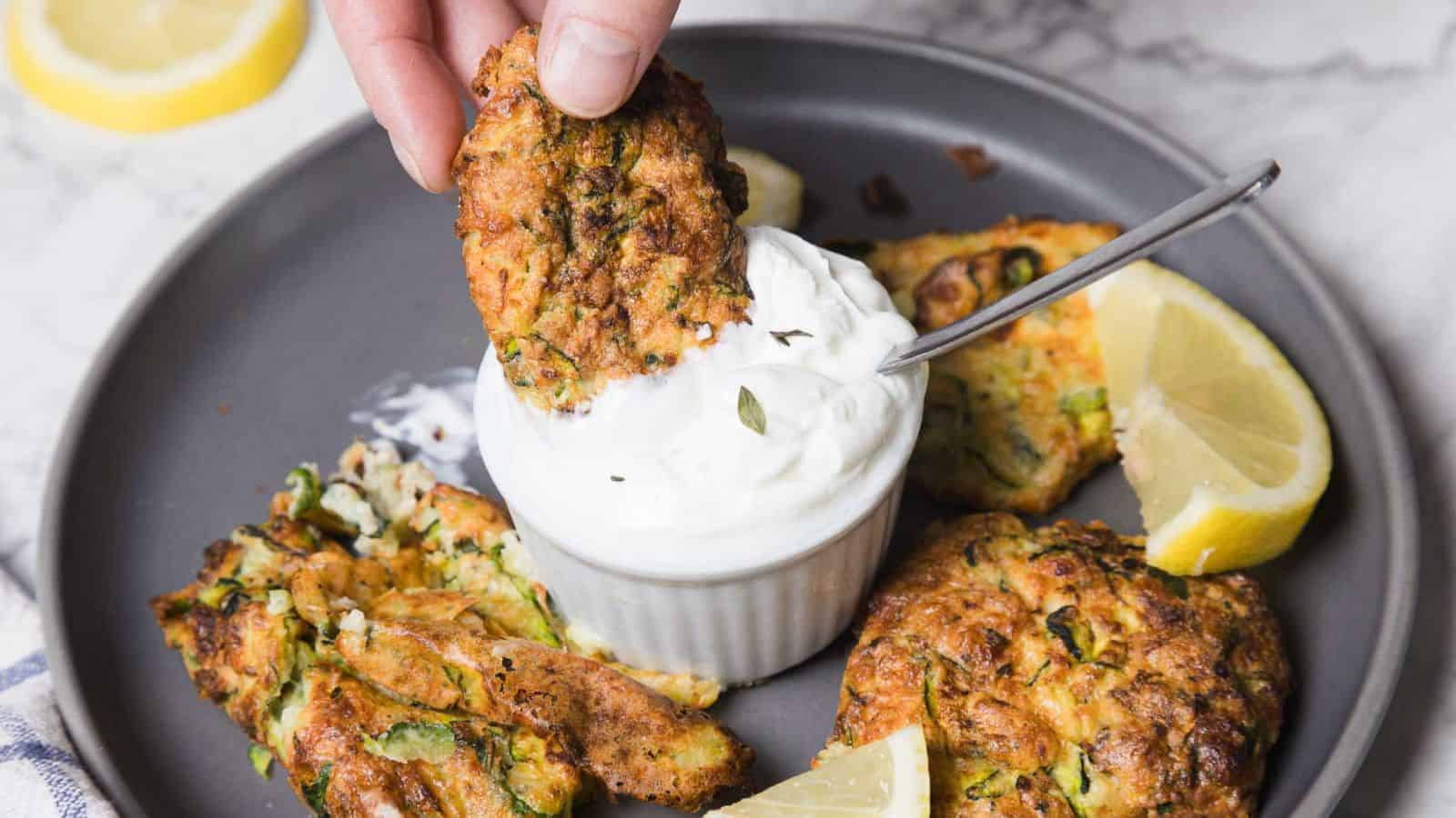A hand dips a zucchini fritter into a white sauce in a small ramekin. The plate contains more fritters and lemon wedges. A spoon rests in the ramekin. The background features a marble surface.