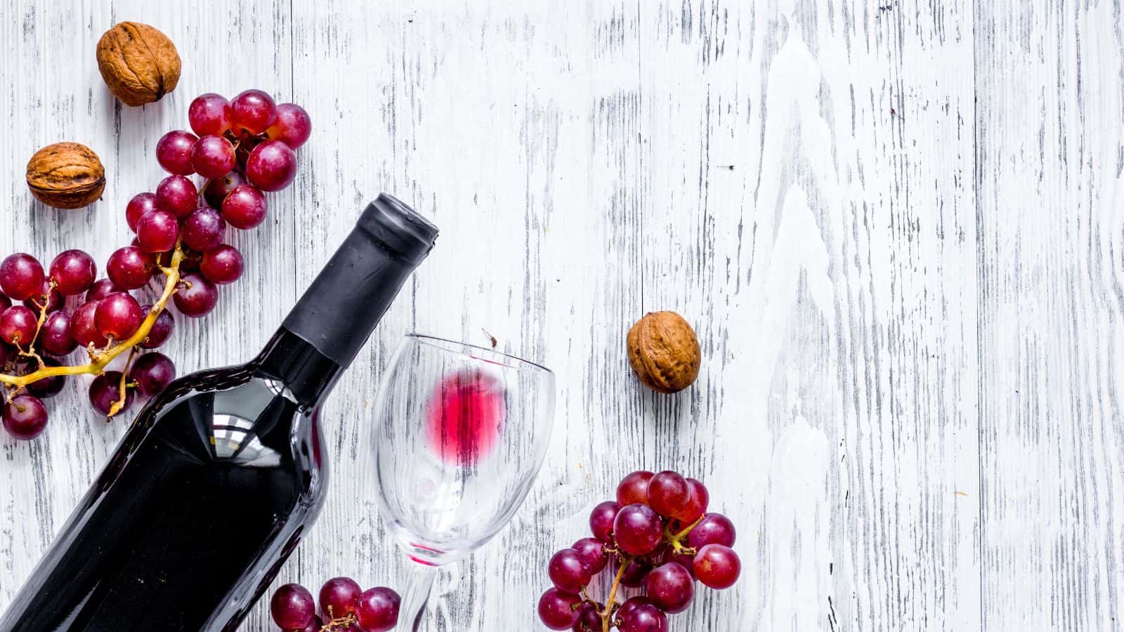 A wine bottle lies on its side next to a partially filled glass with red wine. Surrounding them are clusters of red grapes and a few walnuts. The background is a white, textured wooden surface.
