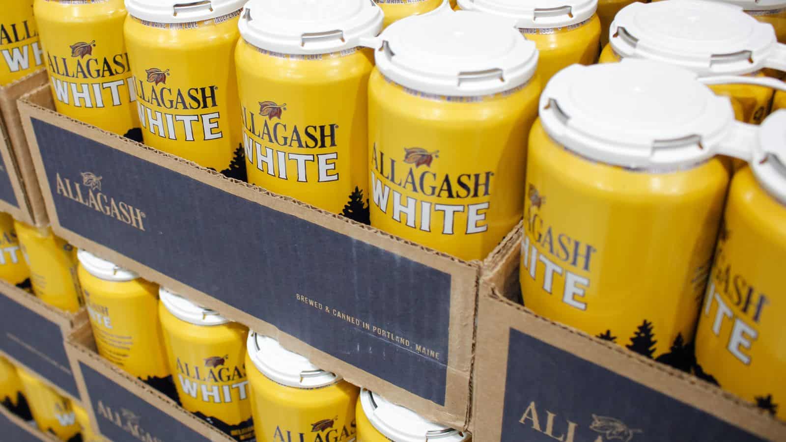 Stacks of yellow cans of Allagash White beer in cardboard trays. The cans are aligned neatly, with the brand name prominently displayed in large letters.