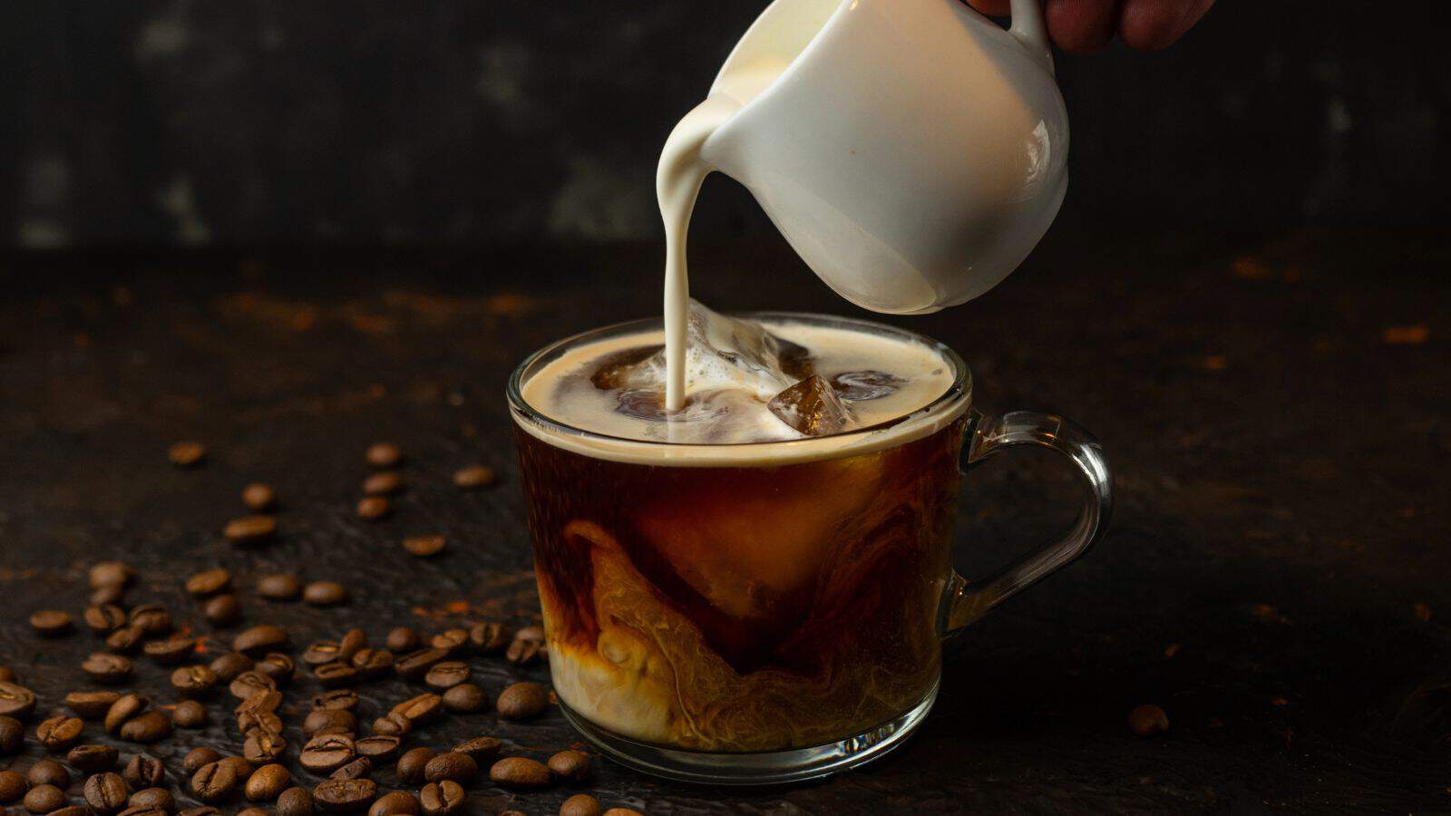 A hand pours cream from a small white pitcher into a transparent glass mug of iced coffee. Coffee beans are scattered on the dark surface around the mug. The background is dark and out of focus.