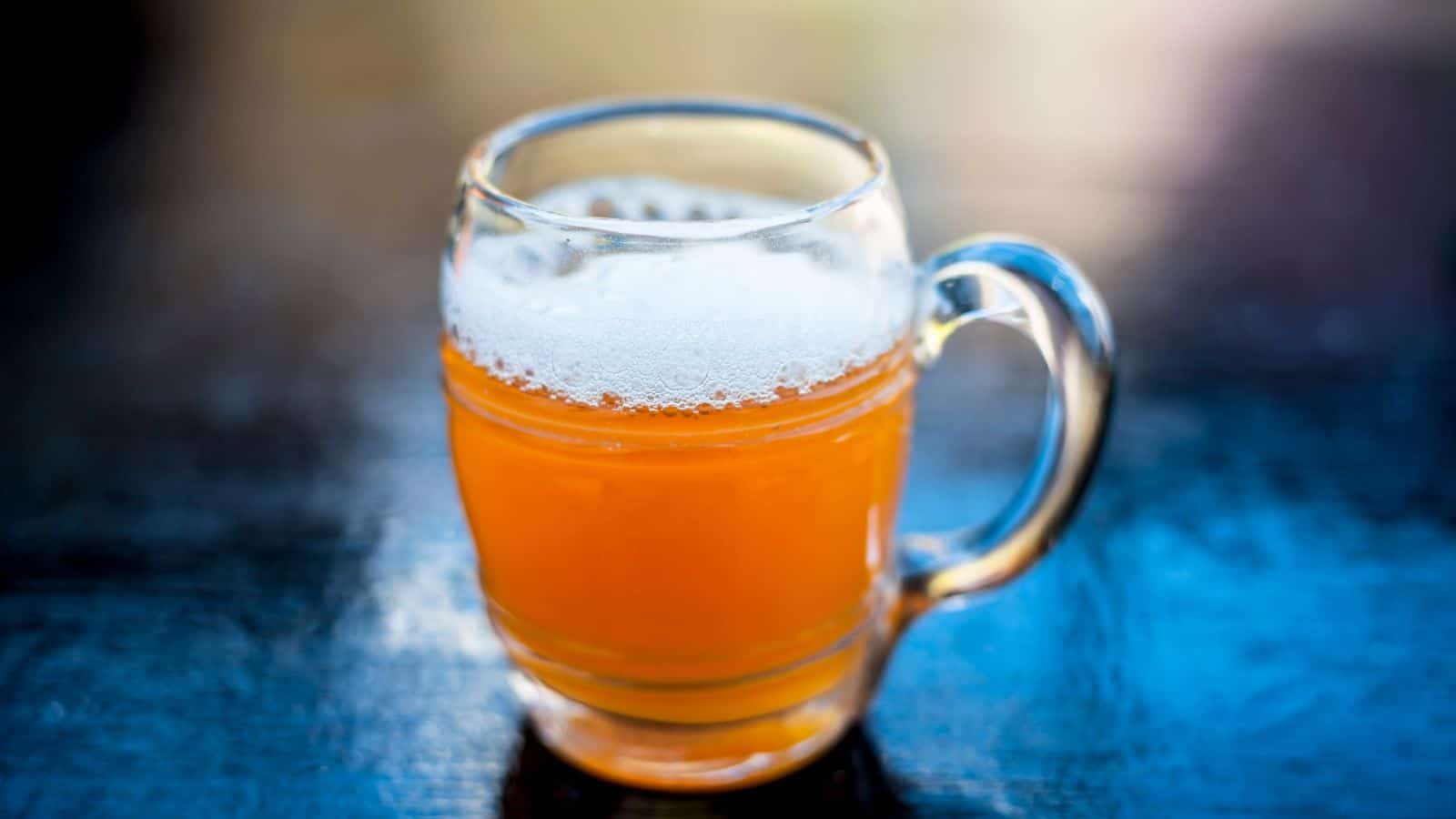 A clear glass mug filled with amber-colored beer, topped with a layer of frothy foam, sits on a dark, reflective surface. The background is softly blurred, highlighting the drink.
