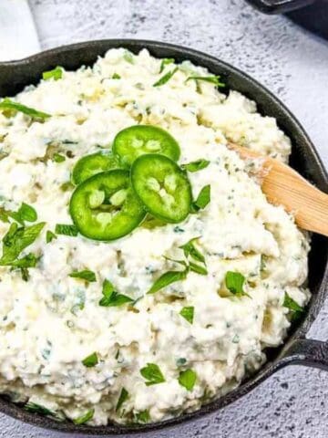 A black bowl filled with creamy dip is topped with sliced jalapeños and chopped herbs. A wooden spoon rests in the bowl. A cloth with lemon designs is partially visible next to it on a textured gray surface.