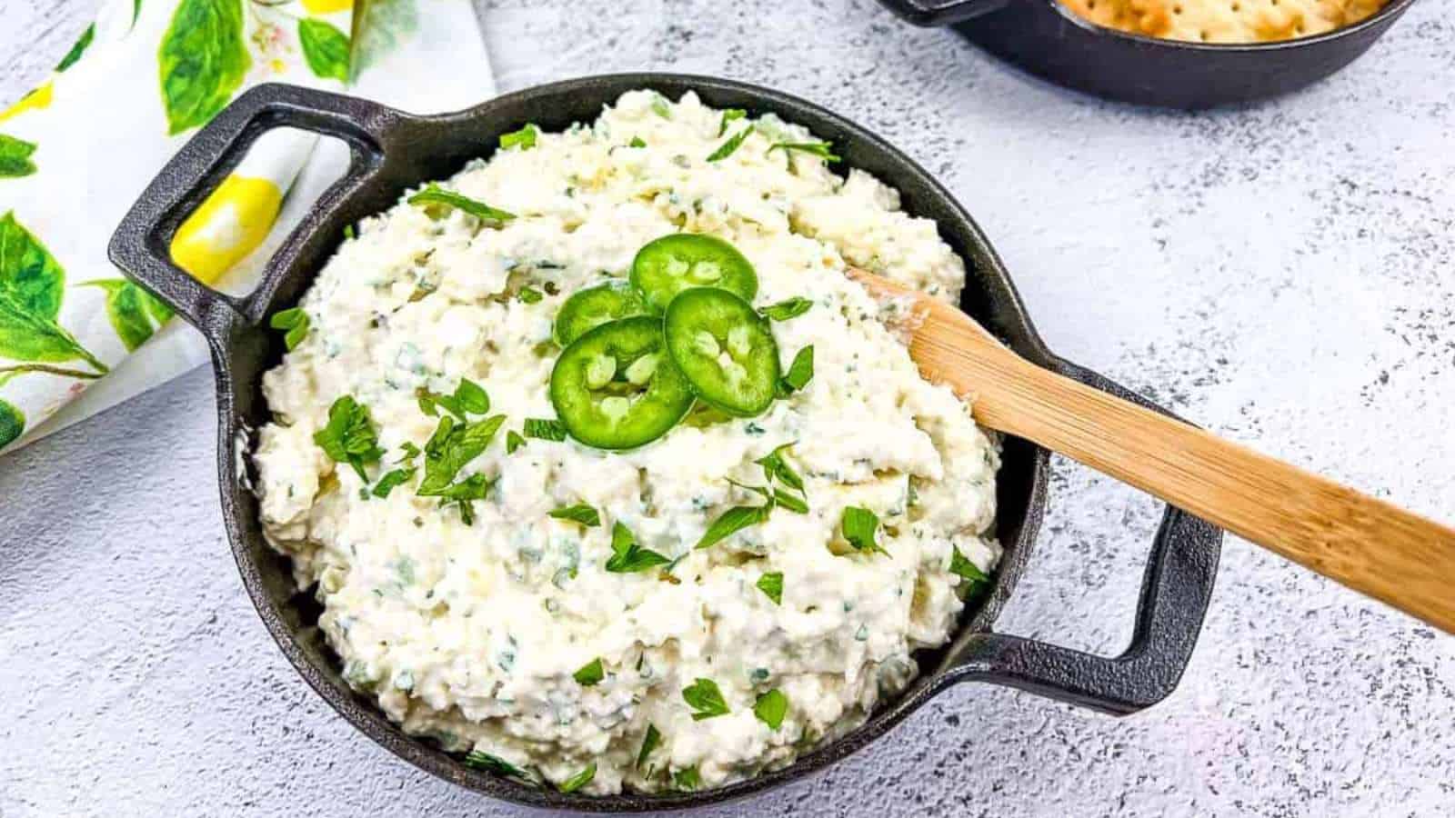 A black bowl filled with creamy dip is topped with sliced jalape&ntilde;os and chopped herbs. A wooden spoon rests in the bowl. A cloth with lemon designs is partially visible next to it on a textured gray surface.