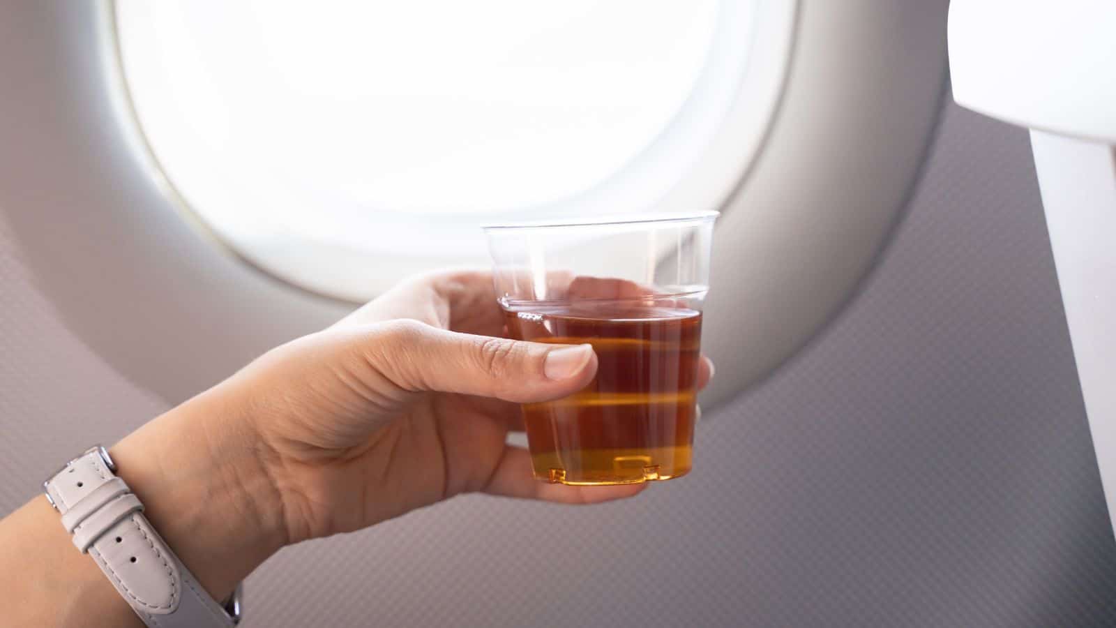 A person holds a transparent cup filled with a brown beverage in front of an airplane window. The person's wristwatch is visible. The window shows daylight entering the cabin.