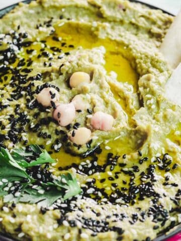 A bowl of green hummus garnished with black sesame seeds, whole chickpeas, a sprig of parsley, and drizzled with olive oil. Two triangular pieces of flatbread rest at the edge of the bowl on a light-colored surface.