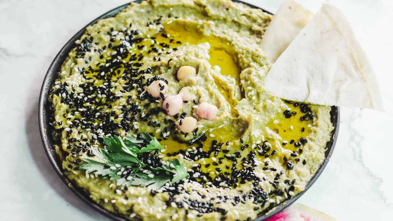 A bowl of green hummus garnished with black sesame seeds, whole chickpeas, a sprig of parsley, and drizzled with olive oil. Two triangular pieces of flatbread rest at the edge of the bowl on a light-colored surface.