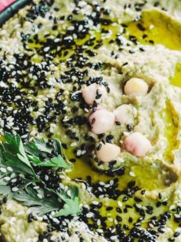 A bowl of hummus topped with black sesame seeds, chickpeas, and a drizzle of olive oil, garnished with a sprig of parsley. Pink and white watermelon radish slices are on the side of the wooden platter.